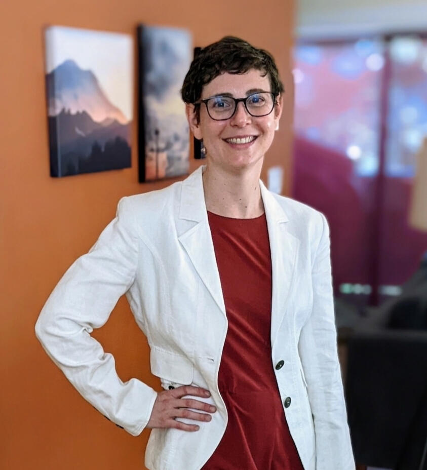 Alison wearing a white blazer and red jumpsuit against an orange wall with two photographs. She is a white woman with short curly brown hair and glasses