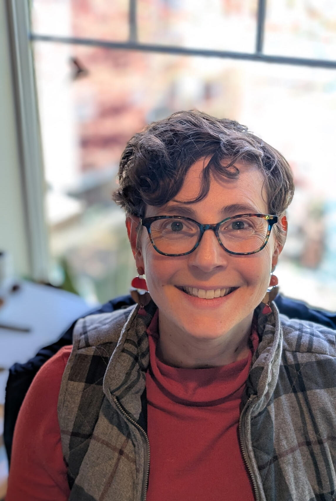 Alison sitting in front of a window with a blurry background. Alison is a white woman with short, curly brown hair and glasses. She is wearing a red orange turtle neck and glasses